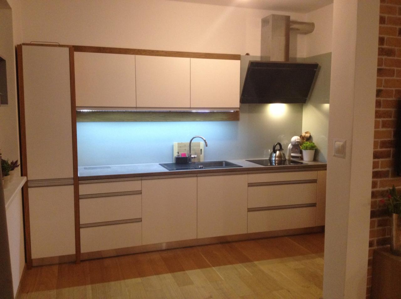 Nowoczesna kuchnia z białymi szafkami i drewnianym wykończeniem, stalowym zlewem i czarnym okapem, oświetlona listwą LED pod szafkami wiszącymi.