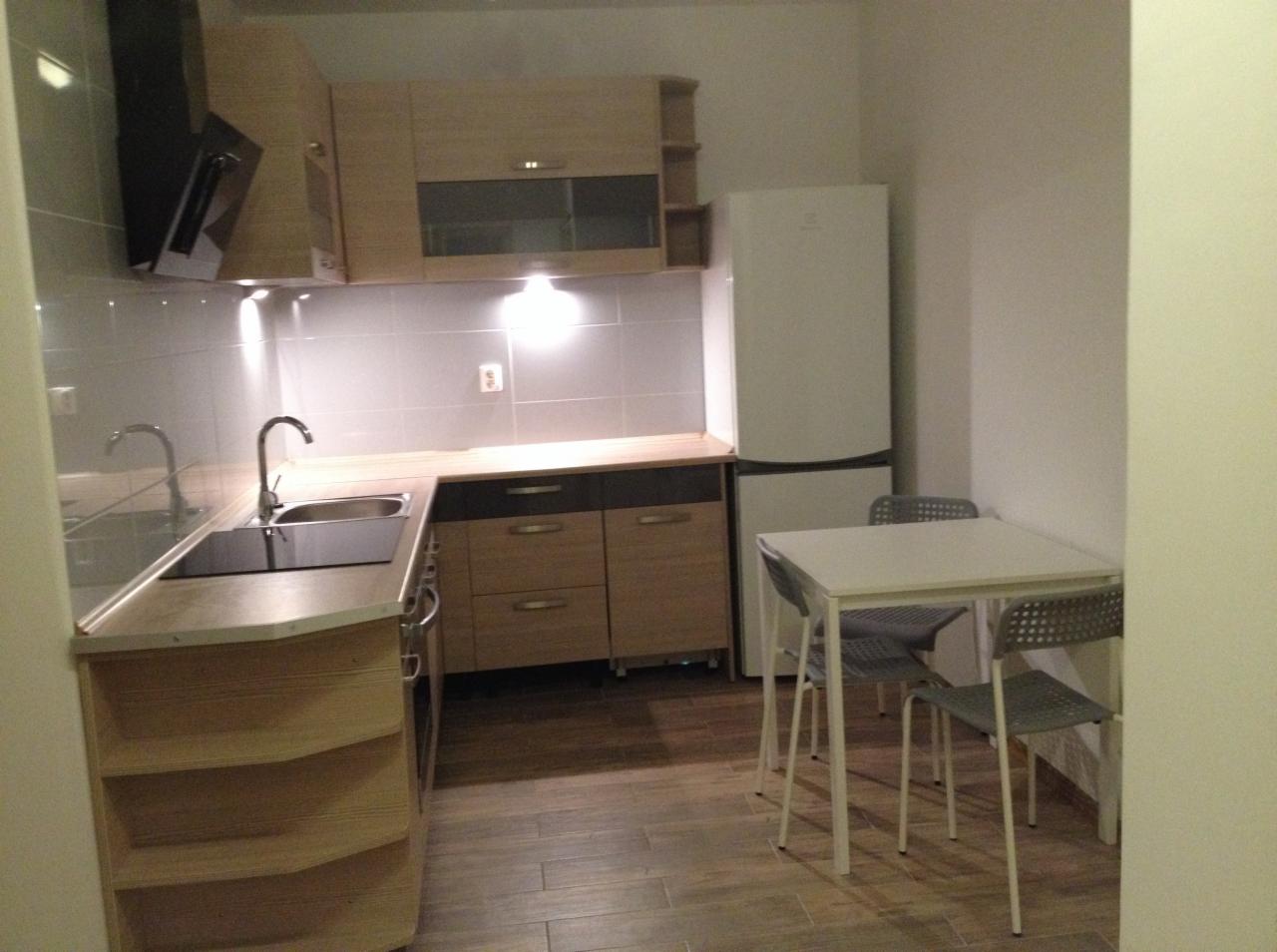 Jasna kuchnia z drewnianymi szafkami, płytkami na ścianie i białym stołem z krzesłami, oświetlona lampkami podszafkowymi.