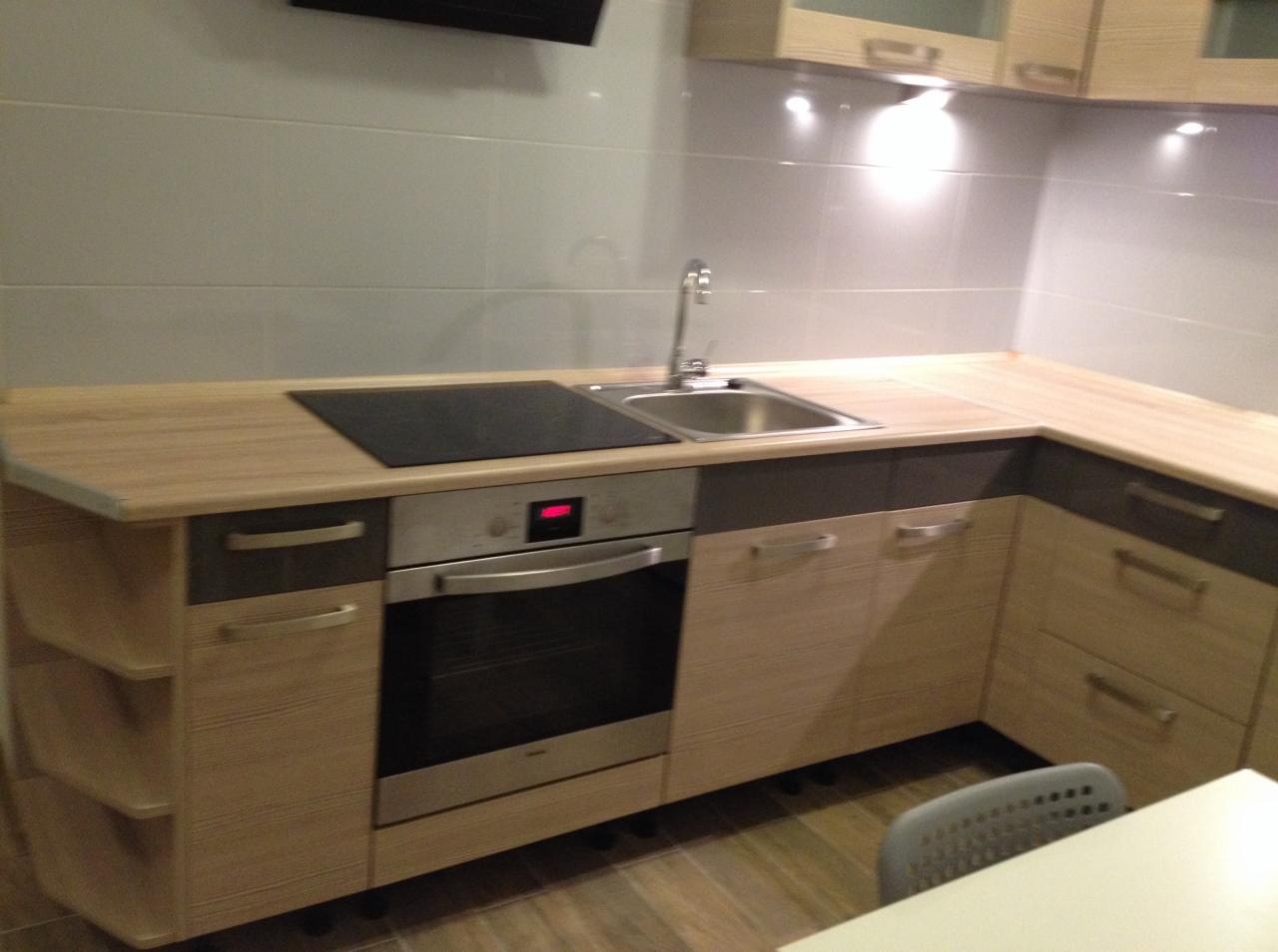 Nowoczesna kuchnia z drewnianymi frontami szafek i blatem, wyposażona w płytę indukcyjną, zlewozmywak ze stali nierdzewnej i piekarnik z wyświetlaczem cyfrowym.