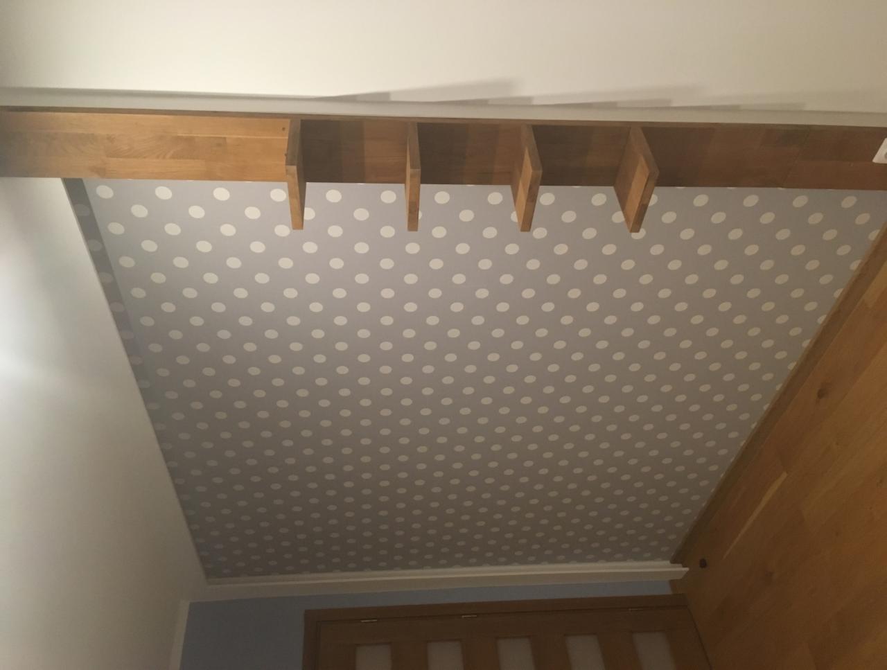Sufit podwieszany z płyt kartonowo-gipsowych wykończony tapetą w białe kropki, z drewnianą belką konstrukcyjną i elementami oświetleniowymi w tle.