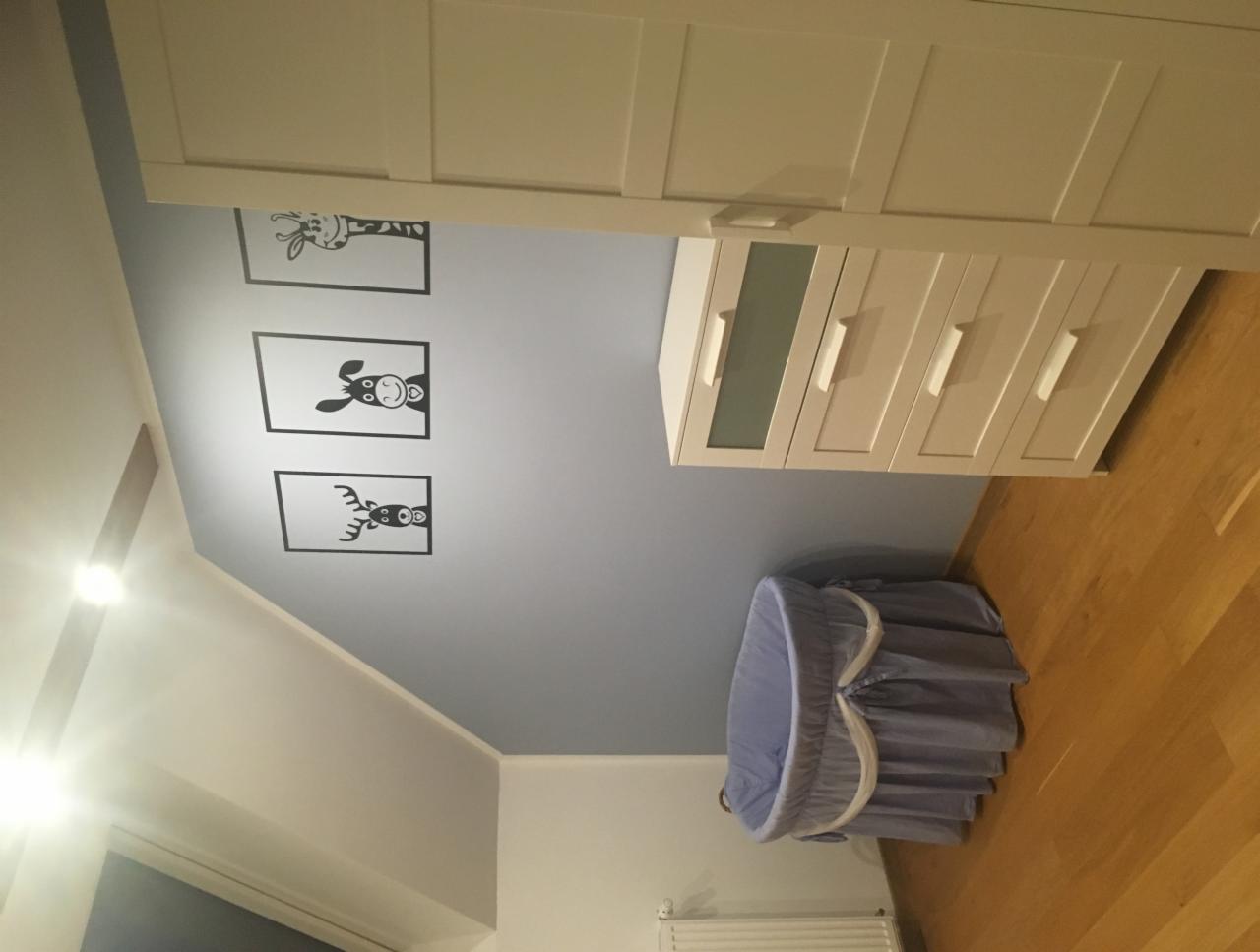 Dziecięcy pokój z niebieskim koszem Mojżesza, białymi meblami i dekoracyjnymi naklejkami zwierząt na ścianie.