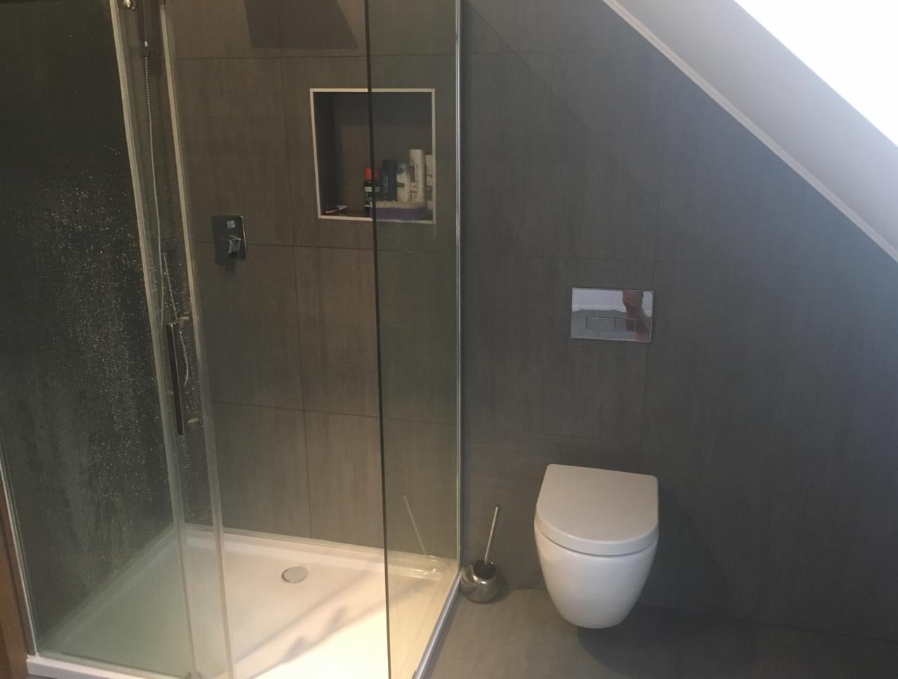 Nowoczesna łazienka w odcieniach szarości z kabiną prysznicową, wiszącą toaletą i wnęką z kosmetykami, zlokalizowana na poddaszu.