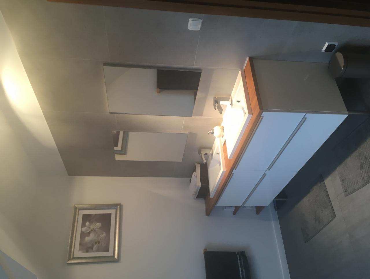 Nowoczesna łazienka z dwoma umywalkami, białymi szafkami z drewnianym blatem, dużymi lustrami i szarą płytką na ścianach oraz podłodze, dekorowana obrazem w srebrnej ramie.