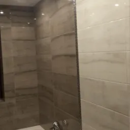 Łazienka wykończona płytkami w odcieniach szarości i bieli, z wanną i armaturą, oświetlona punktowymi lampami sufitowymi, z dekoracyjnym paskiem mozaiki pionowo na styku dwóch kolorów płytek.