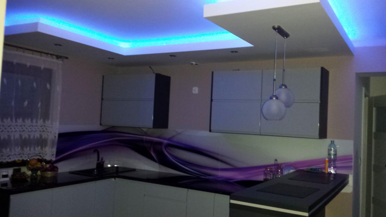 Nowoczesna kuchnia z podświetlanym sufitem LED w kolorze niebieskim, szare szafki wiszące z czarnymi obramowaniami, blat z czarnym zlewem i abstrakcyjnym panelem ściennym w odcieniach fioletu...