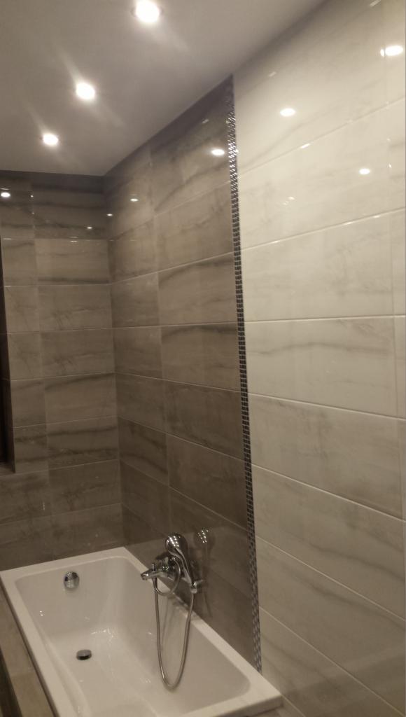 Łazienka wykończona płytkami w odcieniach szarości i bieli, z wanną i armaturą, oświetlona punktowymi lampami sufitowymi, z dekoracyjnym paskiem mozaiki pionowo na styku dwóch kolorów płytek.