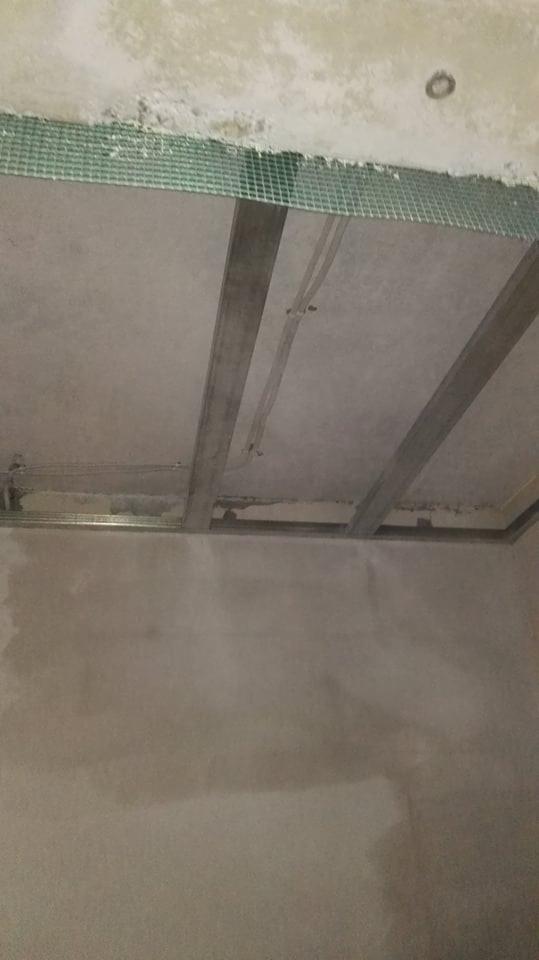 Sufit w trakcie montażu z widoczną metalową konstrukcją nośną i siatką wzmacniającą na obrzeżach, ściany surowe, przygotowane do dalszych prac wykończeniowych.