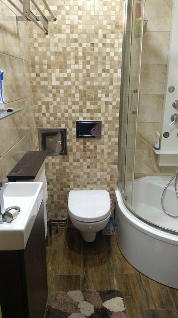 Wykończona łazienka z beżową mozaiką na ścianach, drewnopodobną podłogą, białą umywalką z ciemnym blatem, toaletą i kabiną prysznicową z hydromasażem.