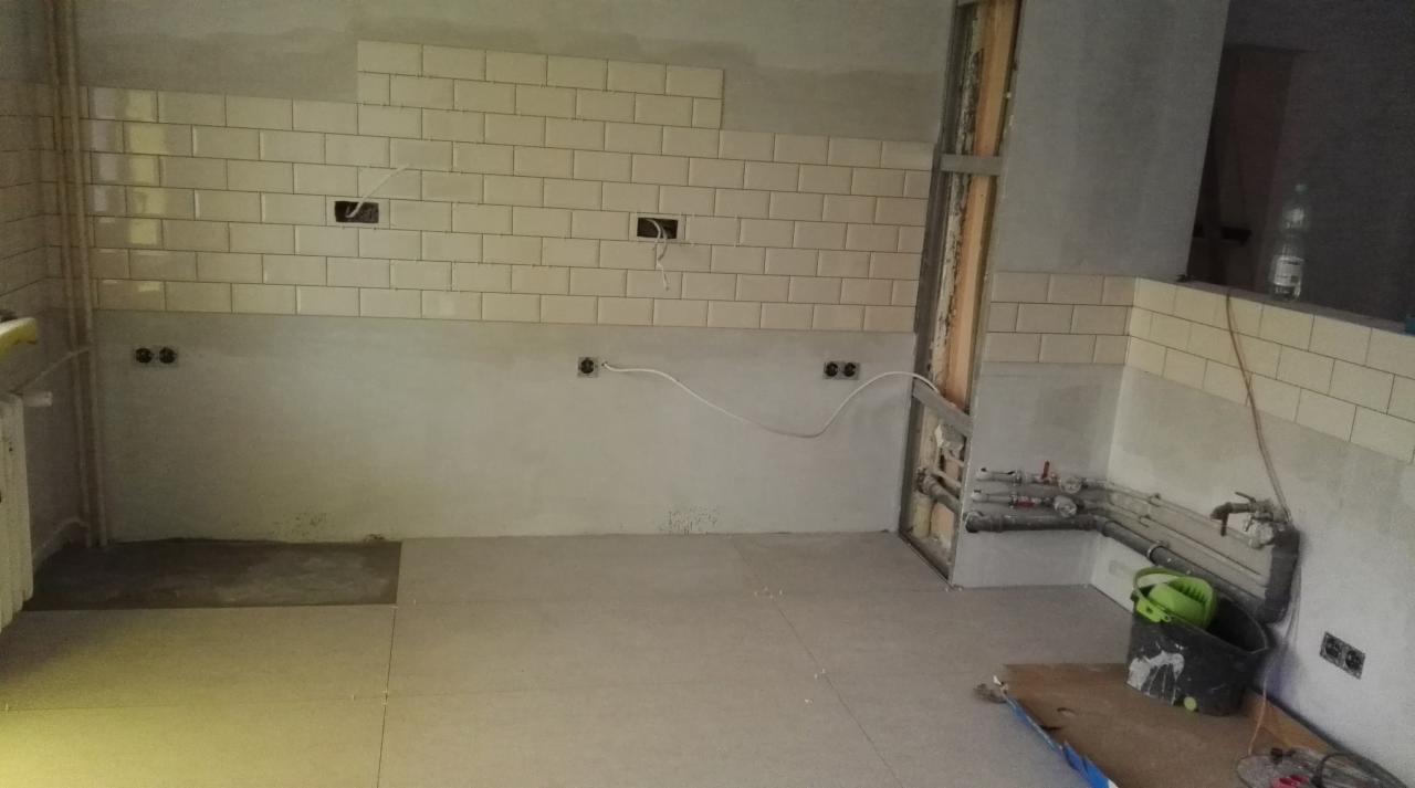Remont kuchni w trakcie: częściowo wyłożone białymi płytkami ściany, widoczne instalacje elektryczne i hydrauliczne, podłoga przygotowana pod dalsze prace, wiadro z narzędziami w rogu.