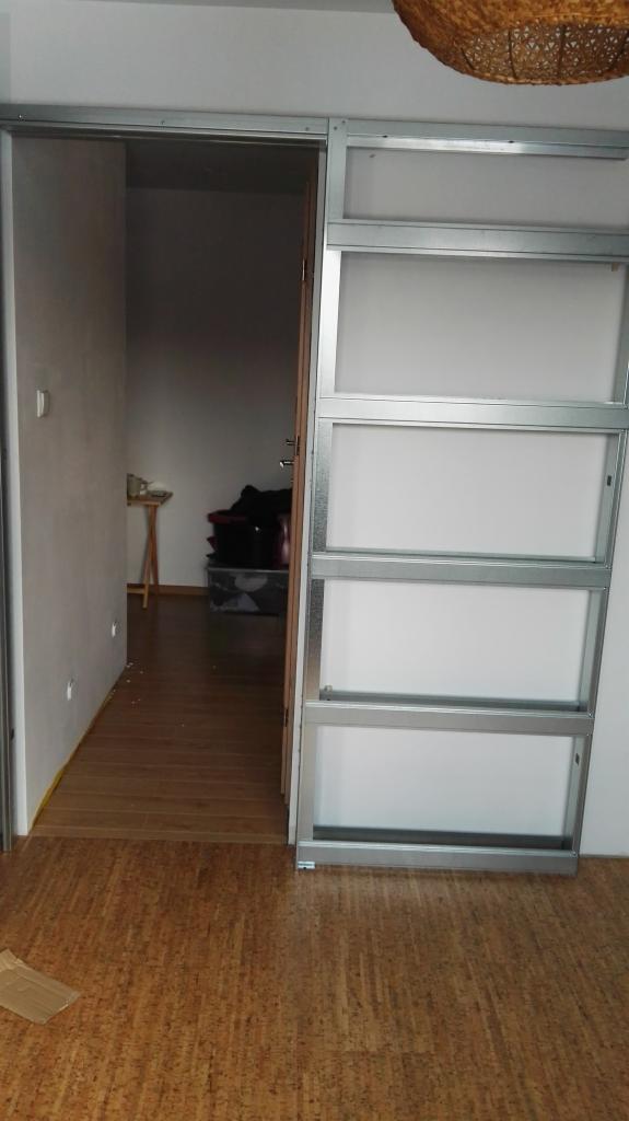 Metalowa konstrukcja ścianki działowej z otwartym przejściem do pokoju z podłogą korkową i widoczną lampą wiszącą z wikliny.