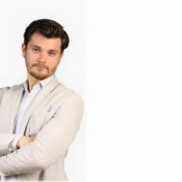 Młody mężczyzna z brodą, w beżowej marynarce i koszuli w kratę, stoi na tle białej ściany z cegieł, z założonymi rękami i spogląda w obiektyw.