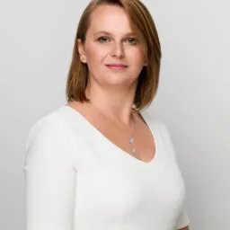 Portret kobiety w białej sukience i srebrnym naszyjniku na jasnoszarym tle.