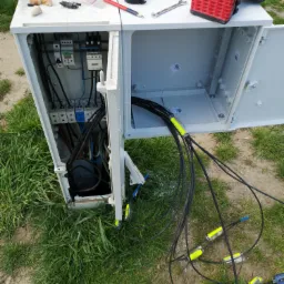 Otwarta szafka telekomunikacyjna z widocznymi kablami światłowodowymi zakończonymi złączami, narzędzia pracy na górze obudowy, trawa w tle.