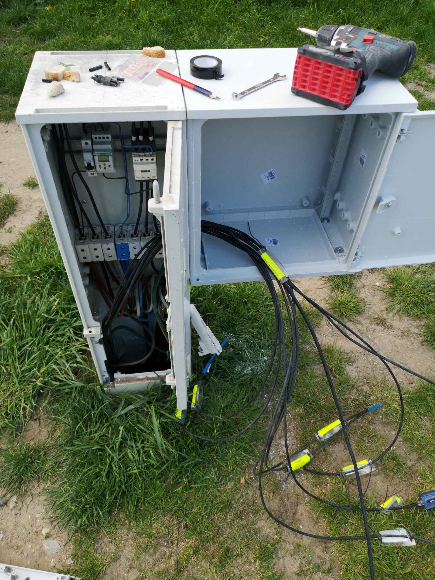 Otwarta szafka telekomunikacyjna z widocznymi kablami światłowodowymi zakończonymi złączami, narzędzia pracy na górze obudowy, trawa w tle.