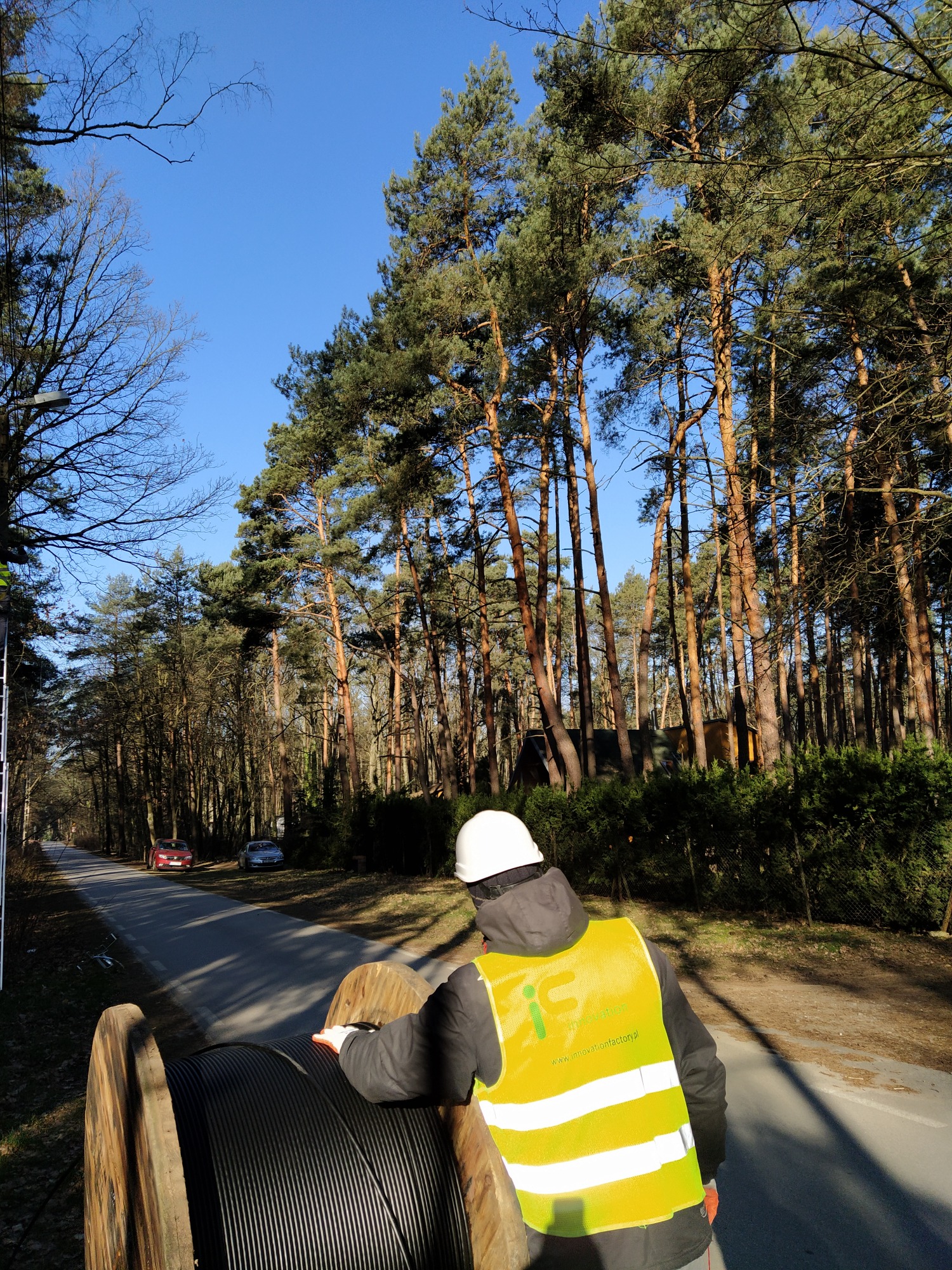 Pracownik w kasku i kamizelce odblaskowej rozwijający kabel telekomunikacyjny z dużej drewnianej szpuli na poboczu drogi, w tle las i dwa samochody.