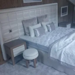 Sypialnia w odcieniach szarości z dużym, tapicerowanym zagłówkiem w kratę, nocną szafką i okrągłym stołkiem obok łóżka.