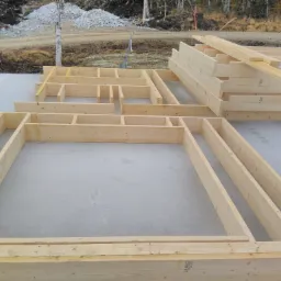 Wstępny etap budowy domu szkieletowego: drewniane ramy ścian ułożone na betonowej płycie fundamentowej, w tle stos belek i teren budowy.