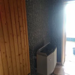 Wnętrze sauny z piecem marki TYLO, wykończone drewnianą boazerią i ciemną cegłą, widoczne niedokończone prace wykończeniowe na podłodze.