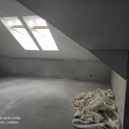 Poddasze w trakcie prac wykończeniowych: zamontowane płyty kartonowo-gipsowe na skosach, dwa okna dachowe, widoczne resztki pianki montażowej na podłodze, drabina oparta o ścianę po lewej.