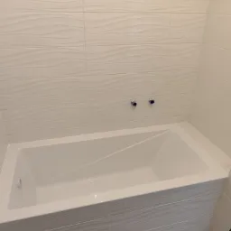 Nowoczesna, biała wanna obudowana płytkami z delikatnym, falistym wzorem, dopasowanymi do płytek ściennych; widoczne niebieskie zaślepki na rurach.