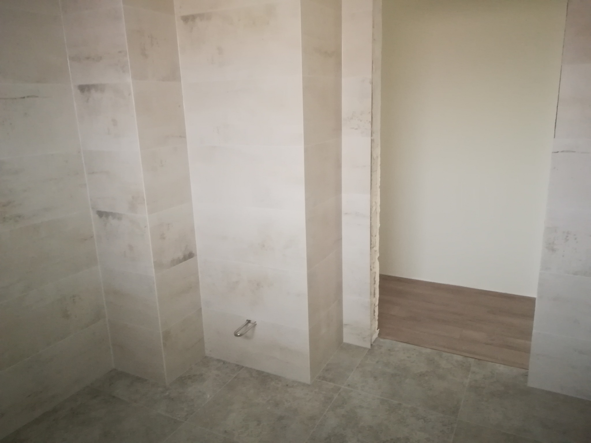 Wykończona łazienka z beżowymi, imitującymi kamień płytkami na ścianach i podłodze; widoczny fragment ściany z zamontowanym uchwytem na ręcznik oraz przejście do pokoju z panelami podłogowymi.