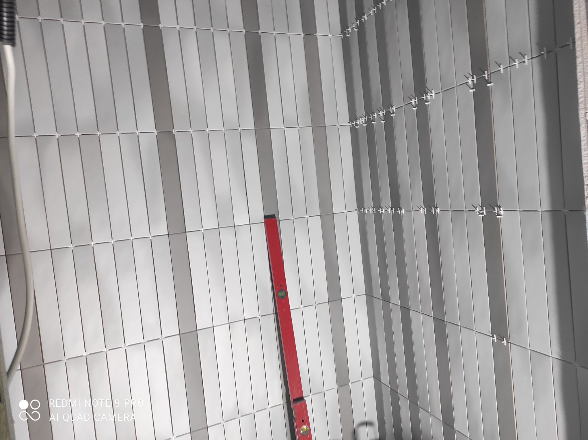 Świeżo ułożone, pionowe, szare płytki w wąskim formacie, z widocznymi krzyżykami dystansowymi i czerwoną poziomicą przy ścianie.