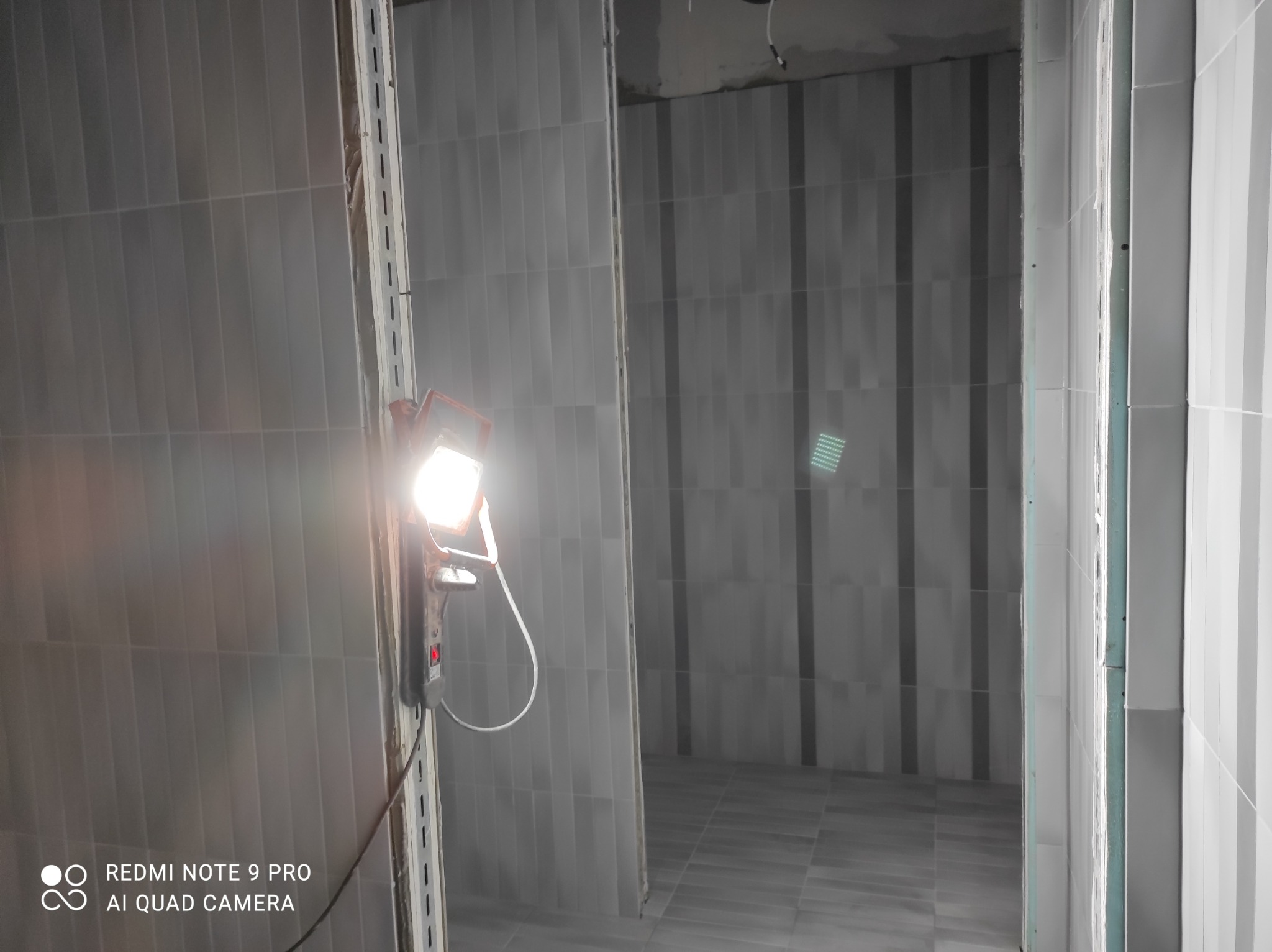 Świeżo ułożone, pionowe, szare płytki w wąskim pomieszczeniu, oświetlone przenośną lampą budowlaną, widoczne niedoskonałości fug.