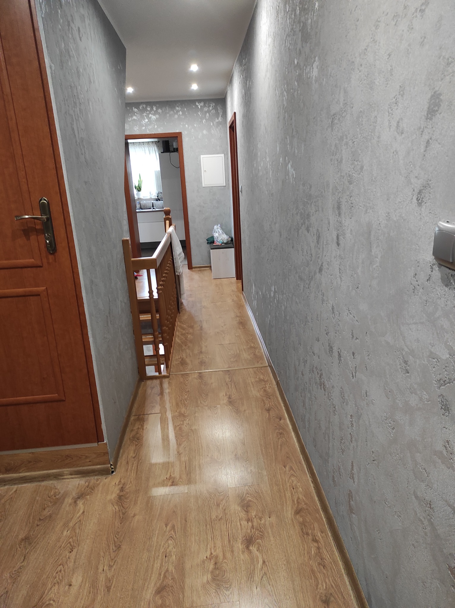 Korytarz z drewnianą podłogą, szarą ścianą o fakturze betonu i drewnianymi drzwiami, widok w głąb mieszkania.
