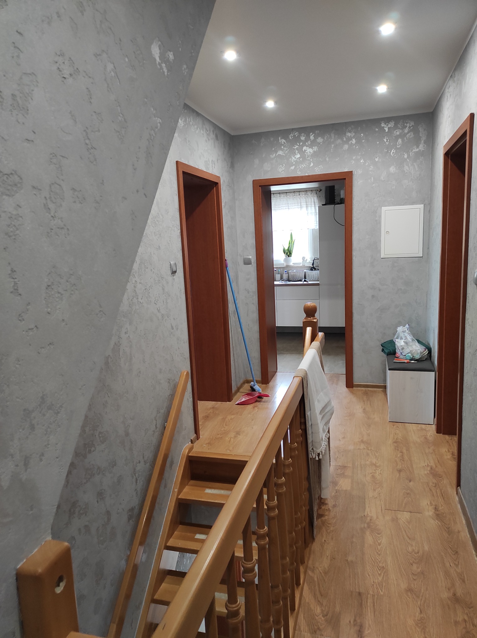 Korytarz z drewnianymi schodami, szarą ścianą o dekoracyjnej fakturze i widokiem na kuchnię przez otwarte drzwi.