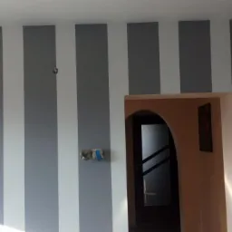 Ściana w szaro-białe pasy, z widoczną instalacją elektryczną w trakcie prac wykończeniowych i fragmentem łukowego wejścia do innego pomieszczenia z obrazem na ścianie.