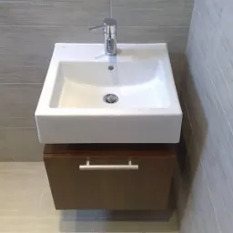 Nowoczesna, kwadratowa umywalka nablatowa z chromowaną baterią, zamontowana na wiszącej szafce z ciemnego drewna, na tle szarych płytek imitujących drewno.