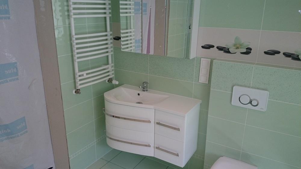 Jasnozielona łazienka z białą umywalką z szafką, grzejnikiem drabinkowym i płytkami z motywem kwiatowym.