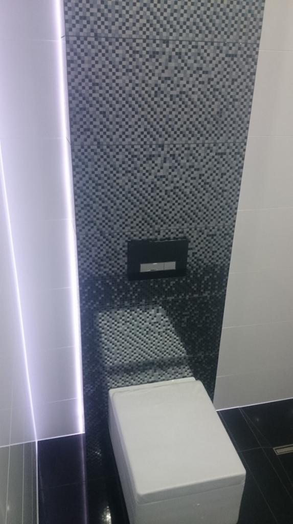 Wisząca toaleta z czarnym przyciskiem spłukującym na ścianie wyłożonej mozaiką w odcieniach szarości, podświetlenie LED wzdłuż ściany.