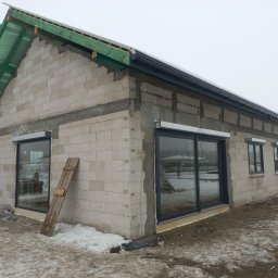 TOKU Projekt - Surowy stan domu z betonu komórkowego z zamontowanymi oknami i przygotowanym dachem, widoczne rusztowania i resztki śniegu na ziemi. Budowa w trakcie.