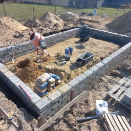 TOKU Projekt - Fundamenty z bloczków betonowych w trakcie budowy, widoczne narzędzia, worki z cementem i betoniarka. Ujęcie z góry, w tle trawa i ogrodzenie.