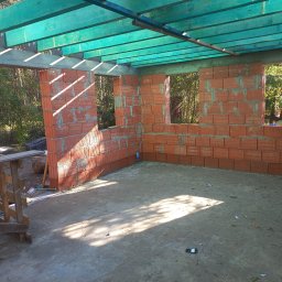 TOKU Projekt - Wnętrze surowego domu z czerwonej cegły z otworami okiennymi i zielonymi belkami stropowymi. Widoczny fragment rusztowania i podłoga w stanie surowym.