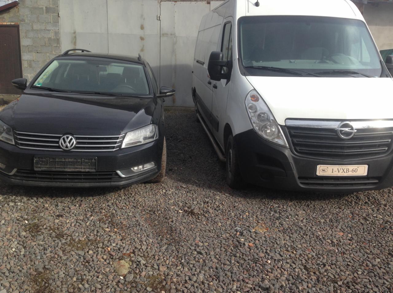 Czarny Volkswagen Passat kombi i biały Opel Movano zaparkowane obok siebie na żwirowym podłożu przed szarą, metalową bramą garażową.