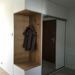 Nowoczesna szafa wnękowa z lustrzanymi drzwiami przesuwnymi i drewnianą wnęką z wieszakami, w której wisi szary płaszcz.