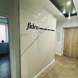Wnętrze kancelarii z marmurową ścianą, na której umieszczono czarne logo 'fideo' i napis 'Kancelaria Radcy Prawnego', widoczne drzwi i fragment podłogi ułożonej w jodełkę.