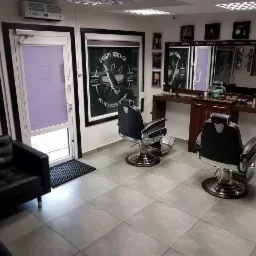 Wnętrze salonu fryzjerskiego z dwoma fotelami barberskimi, czarną sofą i lustrem z oświetleniem nad drewnianą konsolą. Na ścianach grafiki w czarnych ramkach i tablica z napisem 'Fryzjer męski'.