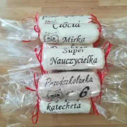 Stos pięciu białych ręczników z czarnymi, wyhaftowanymi napisami (imiona, zawody) i motywami graficznymi (sowy), owiniętych w przezroczystą folię i przewiązanych czerwoną wstążką, ułożonych...