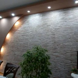 Dekoracyjna ściana z kamienia imitującego piaskowiec, oświetlona punktowymi lampami sufitowymi i dodatkowym oświetleniem wzdłuż łuku, widoczny fragment stołu i krzesła oraz rośliny doniczkowe.