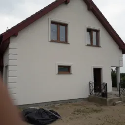 Wykończony dom jednorodzinny z białą elewacją, brązową stolarką okienną i czerwoną dachówką, widoczne elementy dekoracyjne elewacji i balustrada wejściowa.