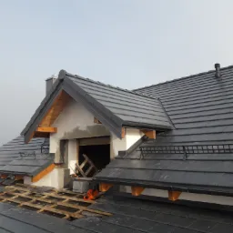 Montaż czarnej dachówki na nowym dachu z lukarną, widoczna więźba i komin, częściowo zdjęte pokrycie, pomarańczowa piła łańcuchowa na dachu.