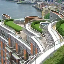 Wielopoziomowy dach budynku mieszkalnego z zielonymi tarasami, trawnikami i basenami, widok z góry na rzekę i zabudowę miejską w tle.