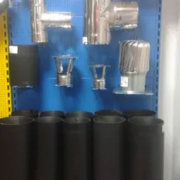 Ekspozycja różnorodnych elementów systemów kominowych: rury stalowe malowane na czarno, kolanka, trójniki i zakończenia kominowe ze stali nierdzewnej na niebieskiej perforowanej ścianie...