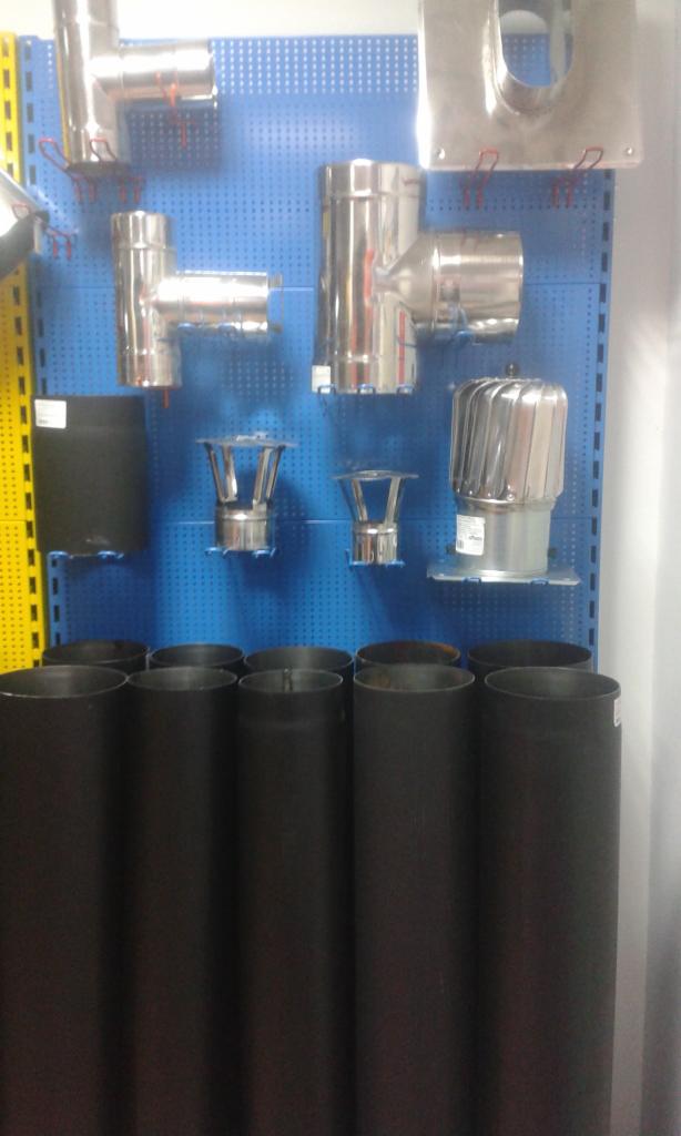 Ekspozycja różnorodnych elementów systemów kominowych: rury stalowe malowane na czarno, kolanka, trójniki i zakończenia kominowe ze stali nierdzewnej na niebieskiej perforowanej ścianie...