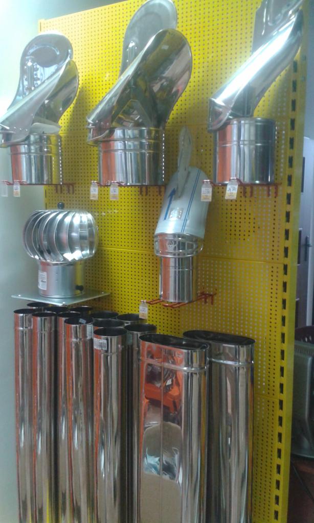 Różne rodzaje stalowych wkładów i nasad kominowych, wyeksponowane na perforowanej, żółtej ścianie w sklepie.