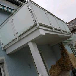 MDJ Metalltechnik Dawid Janczyk - Nowoczesny balkon z balustradą ze szkła matowego i stalowych poręczy, widok z dołu, element elewacji budynku z oknami i roletami.