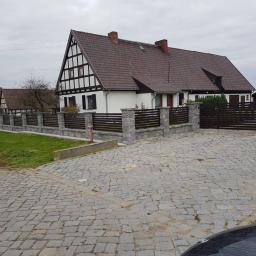 MDJ Metalltechnik Dawid Janczyk - Nowoczesne ogrodzenie posesji z drewnianymi panelami poziomymi i kamiennymi słupkami, uzupełnione bramą wjazdową, na tle domu w stylu szachulcowym pod pochmurnym niebem.